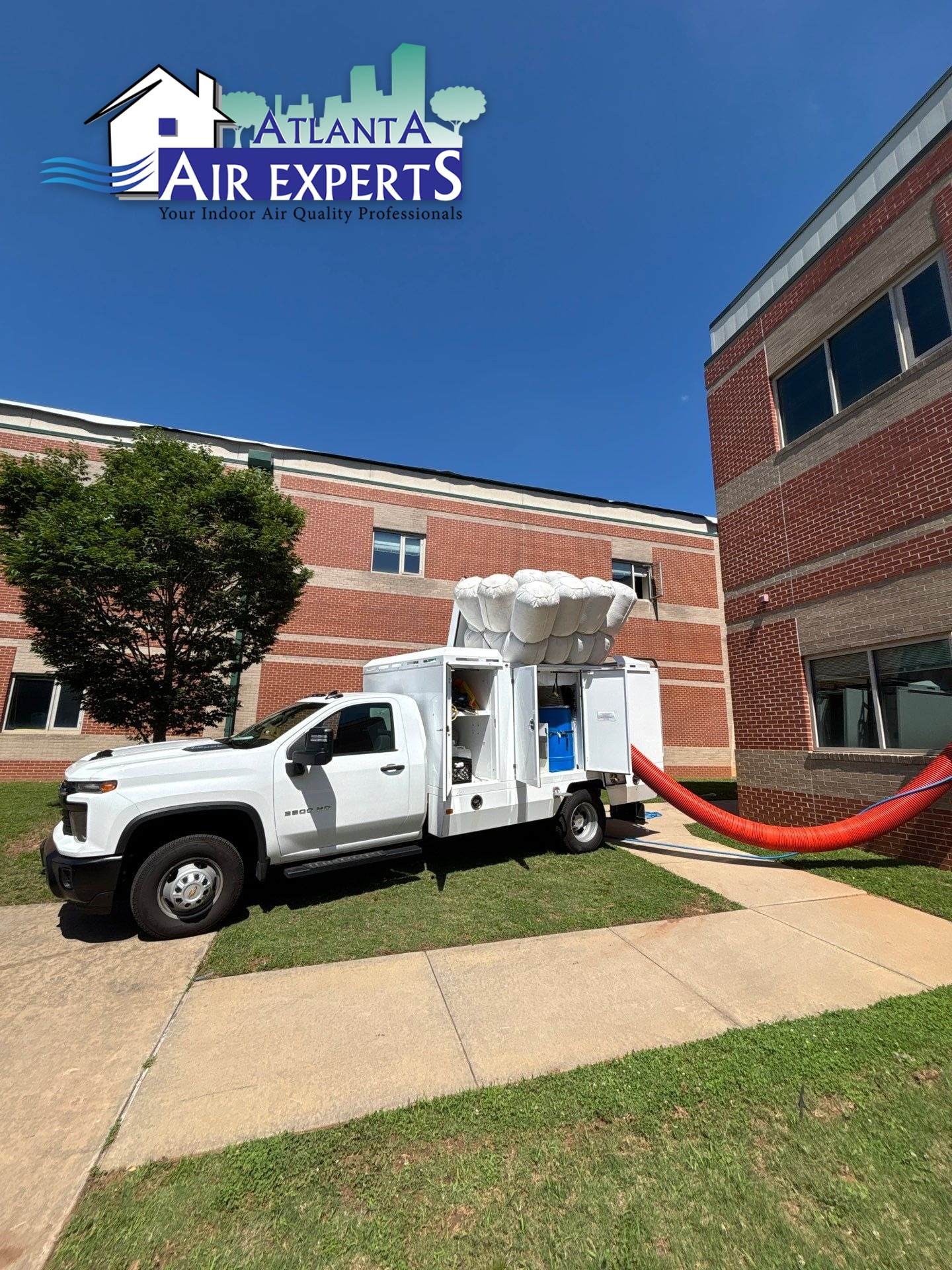 White HVAC service truck with a large duct system on top parked beside a brick office building under a blue sky; logo overlay reads 'Atlanta Air Experts'. The scene conveys air conditioning service operations outside a building.
